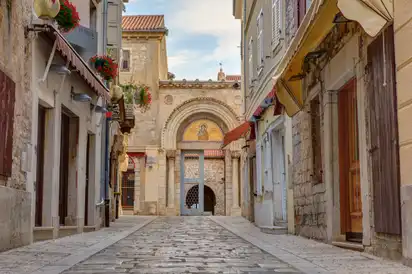 Eine malerische, kopfsteingepflasterte Gasse mit alten Steingebäuden an den Seiten, die zu einer großen, detailliert gestalteten Kirche oder Kathedrale mit einem Bogenportal im Hintergrund führt. Blumentöpfe säumen die Straße, während bunte Markisen den Charme der Szene verstärken.