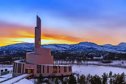 Eine moderne Kirche im Schnee vor einer Bergkulisse während eines bunten Sonnenuntergangs.
