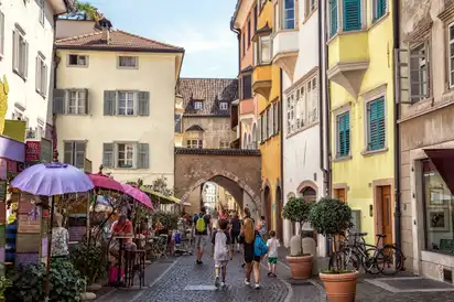 Menschen spazieren durch enge, bunte Altstadtstraße mit Straßencafé.