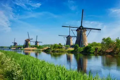 Vier Windmühlen an einem Fluss bei blauem Himmel und grünem Ufer.