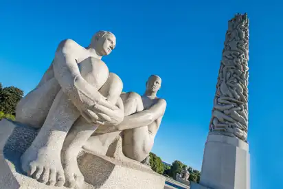 Steinskulpturen von zwei sitzenden Figuren im Vigeland Park, Oslo, mit dem Monolithen im Hintergrund vor klarem blauen Himmel.