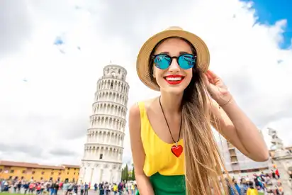 Eine Frau mit Sonnenbrille und Strohhut steht lächelnd vor dem schiefen Turm von Pisa.