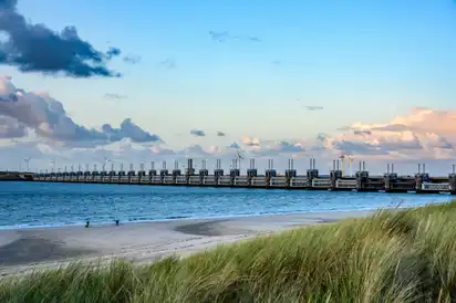 Staudamm an der Küste mit Meer, Sandstrand und Gras im Vordergrund.