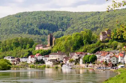 Häuser am Flussufer mit bewaldetem Hügel im Hintergrund, Turm und Burgruine sichtbar.