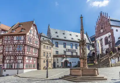 Fachwerkhäuser und eine historische Kirche auf einem gepflasterten Platz unter blauem Himmel.