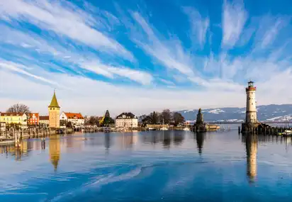 Panorama von Lindau am Bodensee mit dem Hafen und dem bayerischen Löwen sowie dem Leuchtturm im Hintergrund an einem klaren Tag mit blauem Himmel und einigen weißen Wolken.