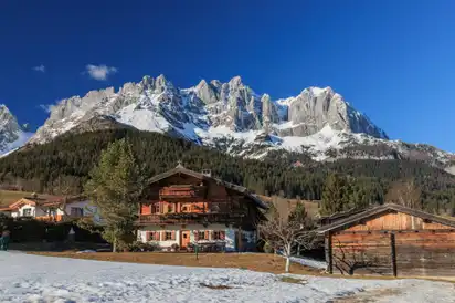 Holzhaus in einer alpinen Landschaft mit schneebedeckten Berggipfeln im Hintergrund und einem klaren blauen Himmel.