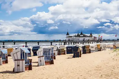 Strandkörbe am Sandstrand vor einer Seebrücke bei bewölktem Himmel.