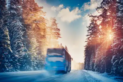 Ein Reisebus fährt im Winter über eine schneebedeckte Straße zwischen verschneiten Bäumen, während die Sonne durch die Bäume scheint.