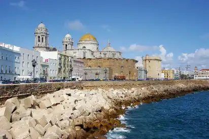 Ansicht der Kathedrale von Cádiz in Spanien mit einer goldenen Kuppel. Im Vordergrund sind die Meereswellen und eine Schutzmauer aus großen, quadratischen Steinen zu sehen. Klare, blaue Himmel und einige umliegende Gebäude sind ebenfalls sichtbar.