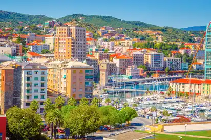 Panoramablick auf die Stadt Savona an der ligurischen Küste mit Gebäuden, Hügeln im Hintergrund und einem Yachthafen im Vordergrund.