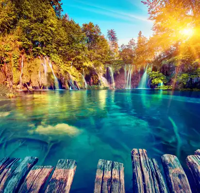 Holzsteg an einem klaren See mit Wasserfällen und Sonnenstrahlen im Wald.