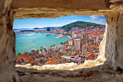 Blick durch Steinfenster auf Küstenstadt mit Meer und Hügel im Hintergrund.
