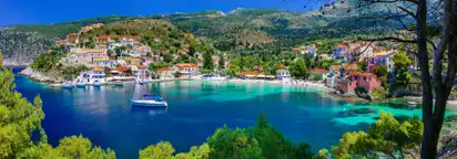 Küstenlandschaft mit griechischem Dorf und Segelboot im türkisblauen Wasser.