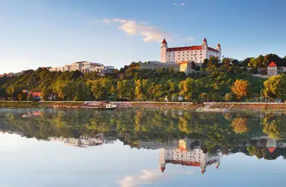 Burg an einem bewaldeten Hügel spiegelt sich im Fluss bei klarem Himmel.