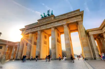 Das Bild zeigt das Brandenburger Tor in Berlin, Deutschland, bei Sonnenuntergang, mit vorbeigehenden Menschen im Vordergrund.