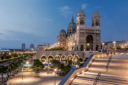 Basilika bei Dämmerung mit beleuchtetem Vorplatz und Treppe in urbaner Umgebung.