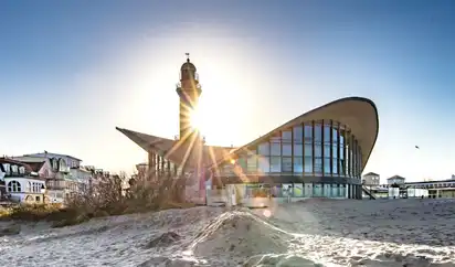 Gebäude des Teepotts Warnemünde bei Sonnenuntergang am Strand der Ostsee in Rostock, Mecklenburg-Vorpommern. Im Hintergrund ist ein Leuchtturm zu sehen.