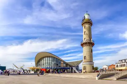Leuchtturm und Teepott in Warnemünde, bei sonnigem Wetter an der Ostsee, mit Menschen am Strand und blauen Himmel im Hintergrund.