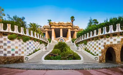 Der Park Güell in Barcelona mit einzigartiger, bunter Architektur und geschwungenen Formen entworfen von Antoni Gaudí, umgeben von Pflanzen und Palmen.