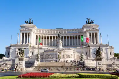 Monumentaler weißer Palast mit Statuen und italienischen Flaggen bei blauem Himmel.