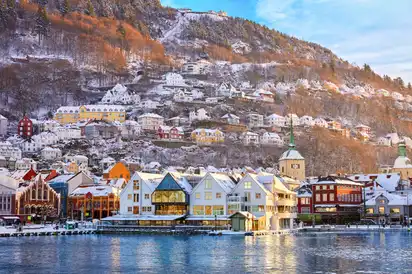 Winterlandschaft mit bunten Häusern am Wasser und schneebedeckten Hügeln im Hintergrund.