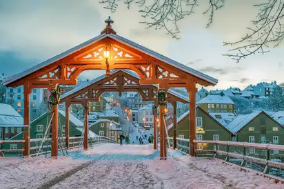 Verschneite Holzbrücke mit Beleuchtung und bunten Häusern im Hintergrund.