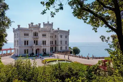Weißes Schloss vor Meer und Sonnenlicht, umgeben von Bäumen.