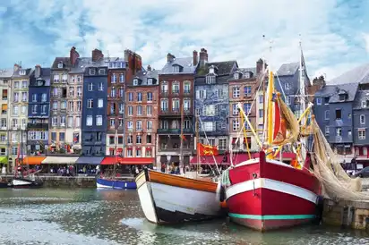 Drei bunte Fischerboote liegen im Hafen vor einer Reihe von charmanten, bunten Häusern im traditionellen Architekturstil. Die Szenerie spiegelt sich im Wasser des Hafens wider, und der Himmel ist mit wenigen Wolken bedeckt.