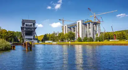 Ein Schiffshebewerk an einem Fluss mit Baukränen und Gebäuden im Hintergrund.