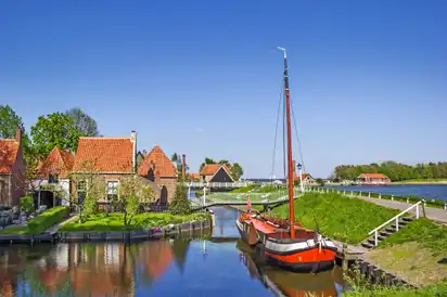Rotes Segelboot in einem Kanal mit Häusern und grünem Ufer unter blauem Himmel.