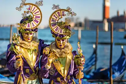 Zwei maskierte Personen in bunten Kostümen vor Gondeln und einem Turm in Venedig.