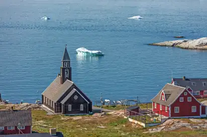 Kirche und rote Häuser an der Küste, im Hintergrund das Meer mit treibenden Eisbergen.