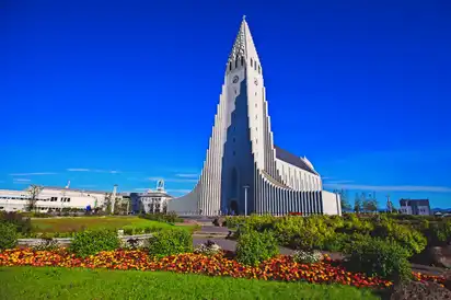 Das Bild zeigt die Hallgrímskirkja Kirche in Reykjavik, Island, mit einem hellblauen Himmel im Hintergrund. Im Vordergrund sind gepflegte Blumenbeete und grüne Rasenflächen zu sehen, die das moderne architektonische Design der Kirche akzentuieren.