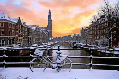 Verschneites Fahrrad auf Brücke vor Kanal und Kirche im Hintergrund bei Sonnenuntergang