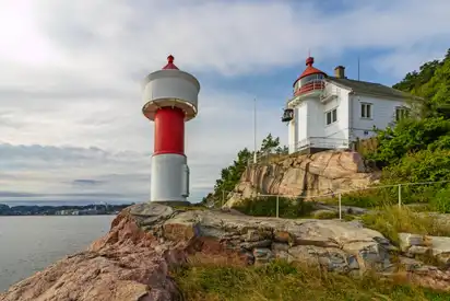 Leuchtturm und weißes Haus auf einem felsigen Abhang mit Blick auf das Meer unter einem bewölkten Himmel.