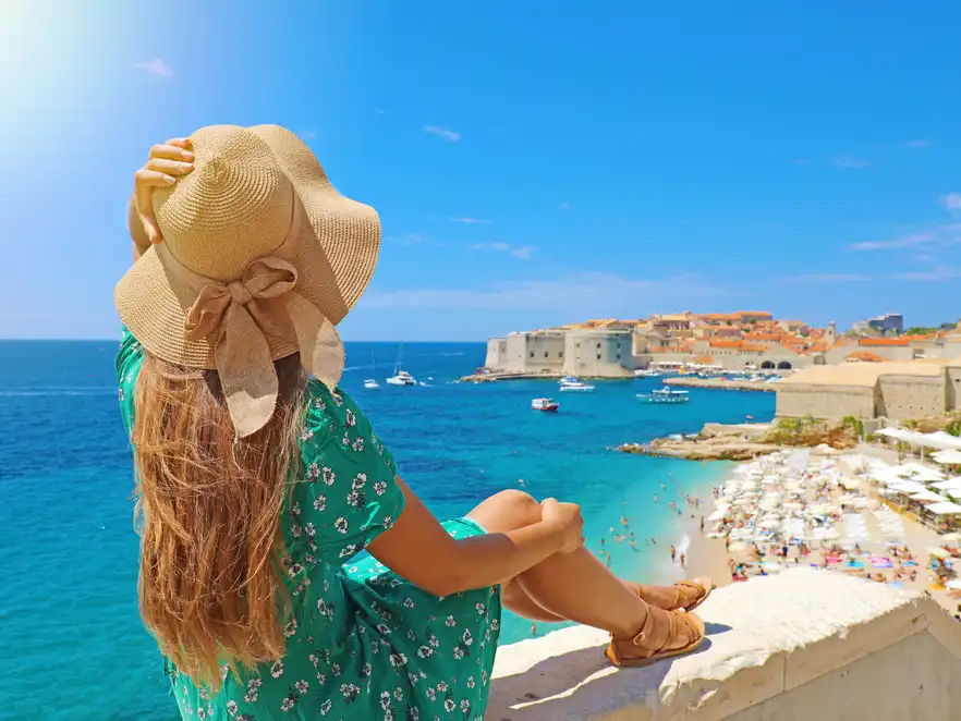 Frau sitzt auf einer Mauer an der Adria und blickt auf die Altstadt von Dubrovnik.